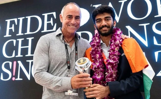 Paddy Upton standing with chess player Gukesh, who is wearing an Indian flag and a garland while holding a trophy